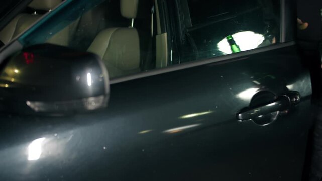 Close view of drunk driver unlocking black vehicle door. Adult man in grey cloth holding green bottle with alcohol while tryes to open car door. A man is about to drive while intoxicated at night