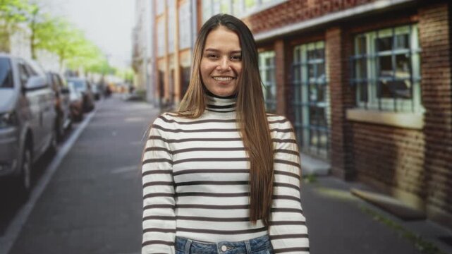 Woman giving two thumbs up with visible thumbs toward camera on a street beside brick building and parked cars, smiling in a striped turtleneck and jeans; confidence.