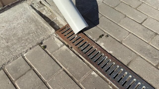 White downpipe and metal grate on paving stones, sunlight casting shadow across textured pavers, closeup view of storm water