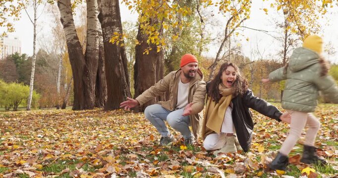 Family parents mother father child autumn park running. Smiling adults crouch with arms wide as a girl runs through leaves, enjoying outdoor fun and quality time. Family bonding theme for ads.