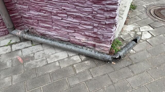 Cracked pipe by brick corner leaking onto pavement, corroded metal downspout detached from wall, small hole releasing water, moss