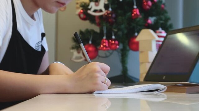 사람 프리랜서 업무 필기 노트북 작업 오피스 인테리어 소품 일상 디테일 자재: 실내 작업실에서 크리스마스트리를 배경으로 노트북을 켜고 노트에 필기하는 인물의 모습을 포착한 바스트숏입니다.