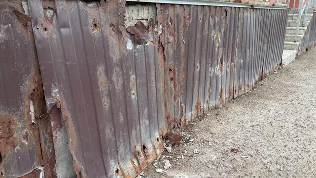 Rusty corrugated metal cladding on extension, close-up view of peeling paint and heavy corrosion along base. Gravel foreground