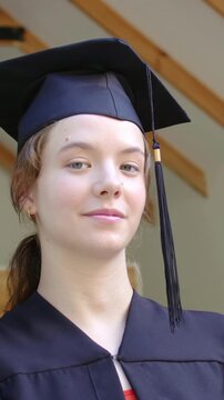 Vertical video: Smiling grad reacting to cam, tilting head, looking at lens under porch, cap and gow