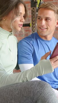 Vertical video: Raising woman in green gym top showing pink phone to man on gym bench, he smiling