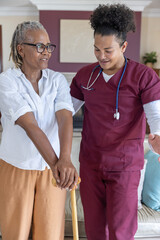 Obraz premium Senior African American woman holding cane, nurse with stethoscope guiding her in living room