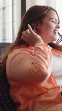 Vertical video: Tucking hair, woman gesturing settling by window for camera, peach shirt