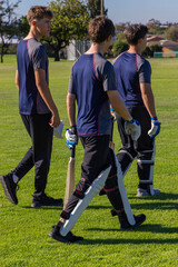 Obraz premium Diverse teen male cricket players walking across field holding bats and wearing navy shirts