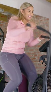Vertical video: Gripping handles woman pedaling airbike at home for fitness raising arm, pink top