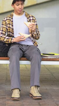 Vertical video: Inspecting snack, unwrapping student in beanie plaid on bench with backpack to eat