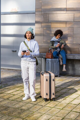 Female friends waiting by stone bench outside wearing beanie, holding smartphone and suitcases © wavebreak3