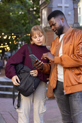 Obraz premium Male and female friends checking smartphone at plaza by stone monument under string lights