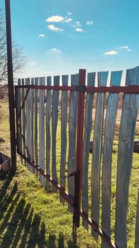 Weathered metal gate with vertical stainless strips, chain link fences flanking sides, spring grass carpeting ground, clear blue