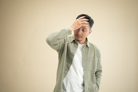 Stressed young Indonesian man doing facepalm gesture with hand on forehead against beige studio background.