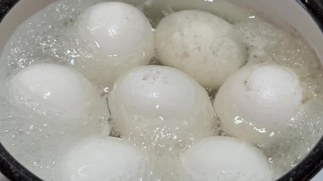 Seven white eggs boiling in ladle over gas stove close-up, enamel ladle filled with bubbling water and rising steam, glossy shells