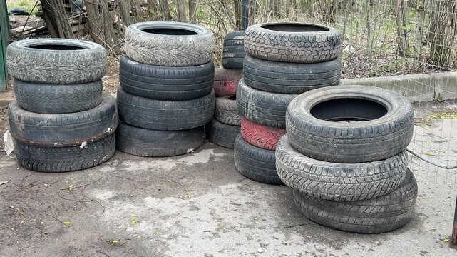Stacked worn tires in illegal dump on street curb beside rusted fence and gravel, muddy pavement speckled with moss and red paint