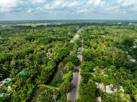 Aerial view of a long asphalt road cutting through a dense tropical forest with a parallel canal and lush green palm trees under a cloudy sky Bangladesh.