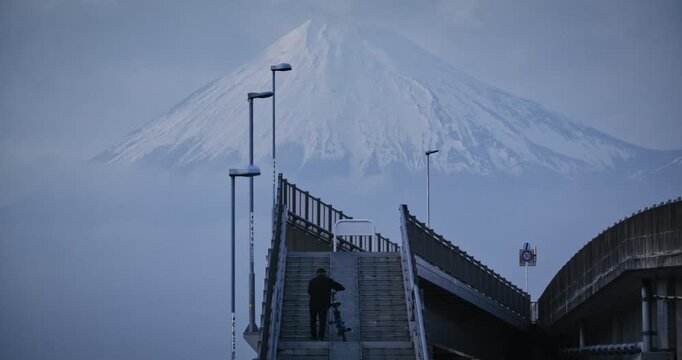 Silhouette of person with bike walking up steps of bridge with beautiful view of hazy Mt. fuji in background at dusk