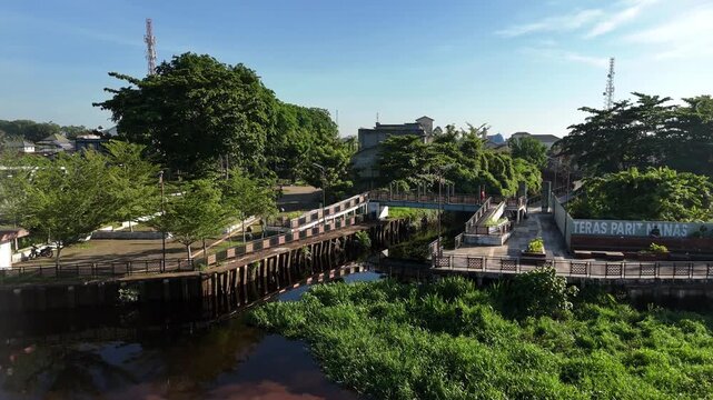 A Path and Pedestrian Bridge in Riverside Park Crossing an Urban Canal on a Sunny Morning at Teras Nanas in Pontianak, Indonesia.