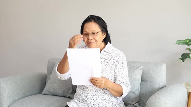 Stressed frustrated Asian older woman getting bad news from paper letter, reading document at home, touching head in despair, panic attack, sitting on couch, receiving concerning medical result