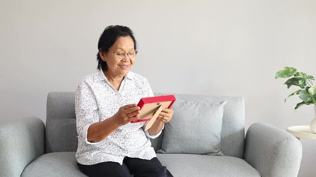 Asian Senior woman looks family photographs in living room. Loving beautiful elderly older female grandmother sitting on sofa enjoy retirement life, missing husband and relative in house.