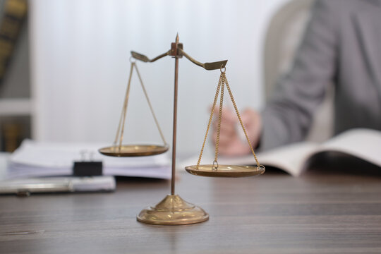 A pair of vintage golden scales of justice rests on a wooden desk with a blurred figure of a lawyer and a book in the background.
