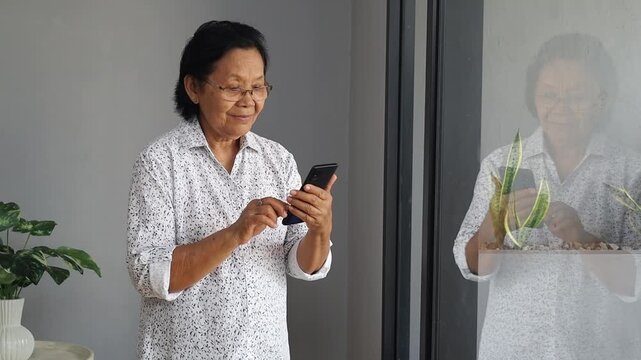Happy senior Asian woman standing by window, holding cellphone in hands, elderly lady contacts her family