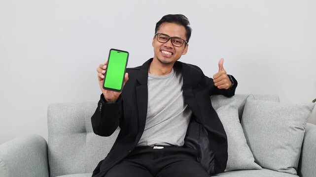 Asian man at home, sitting on the couch and showing the camera a green mock-up of a mobile phone and a super finger sign.