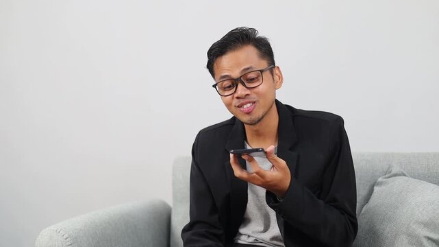 Asian man sitting on sofa at home and talking on speaker phone, recording voice message, dictating his conversation on mobile app
