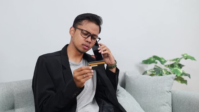 Asian businessman talking on phone and holding a credit card, experiencing financial stress and frustration trying to resolve a serious payment issue