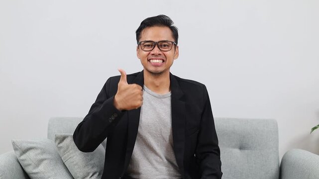Asian male adult businessman wearing suit and glasses, sitting on living room sofa, smiling happily while showing thumbs up looking at camera, giving review, support, agreeing or liking something.
