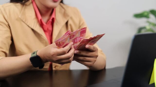 Close up of a businesswoman's hands sitting at her office desk with a laptop on the table, counting rupiah banknotes then fanning them, the concept of getting profit or income from her business.
