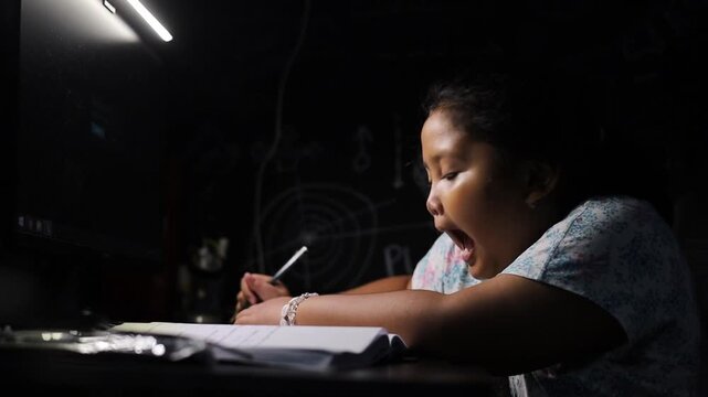 A little elementary school girl is learning to count with her fingers, doing math homework, sitting at a desk with a book and writing with a pencil.