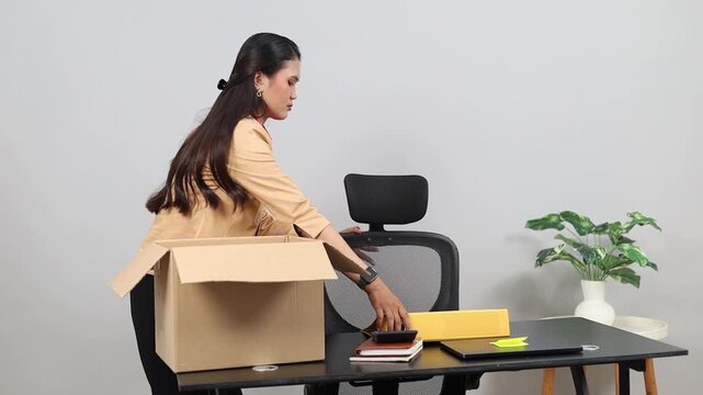 A female worker is in an office space tidying up things on her desk, putting them into a brown cardboard box, with a sad expression, the concept of layoffs or resignation.