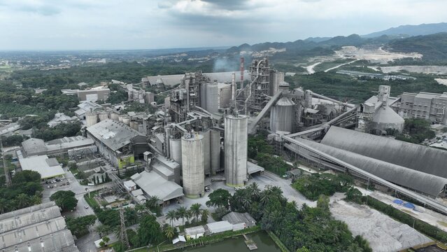 Aerial view of a large industrial factory complex with tall silos, chimneys, and conveyor belts surrounded by lush green trees and hills in Klapanunggal, West Java, Indonesia.