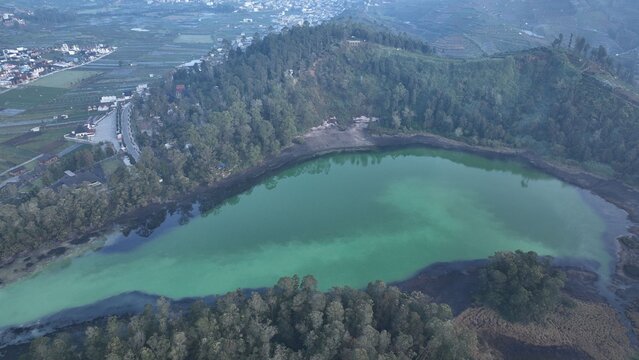 Aerial view of Dieng volcanic lake with turquoise water surrounded by lush green hills and forest in Kejajar, Central Java, Indonesia.