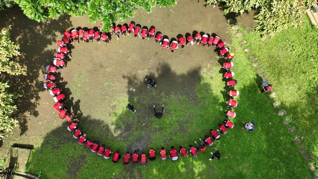 Aerial view of a group of people in red shirts forming a large heart shape on a clearing surrounded by lush green trees in Pangandaran, West Java, Indonesia.