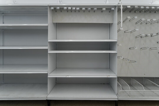 Unstocked white metal grocery store shelving unit standing next to an empty retail pegboard stand with hanging metal hooks.