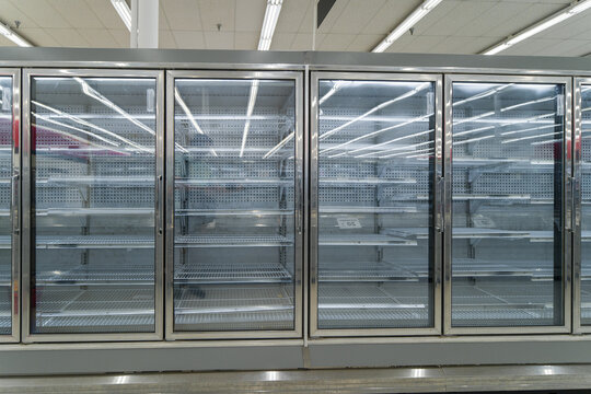 Row of completely empty commercial supermarket glass door refrigerator cabinets standing inside an unstocked retail grocery store.