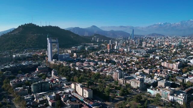 Advancing flight over low density residential neighborhoods approaching San Cristobal Hill with full city skyline and snow-capped Andes in background. Santiago, Chile