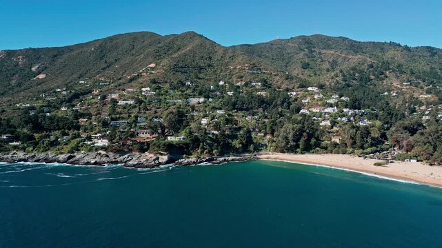 Descending shot over deep blue bay toward rocky coastline with exclusive villas hidden among native forest cascading down to cres