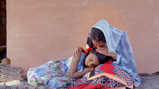 Village scene of a beautiful Indian woman putting her little daughter to a peaceful sleep. Medium shot of a cute little girl sleeping in her beautiful mother's lap during daytime - village family l...