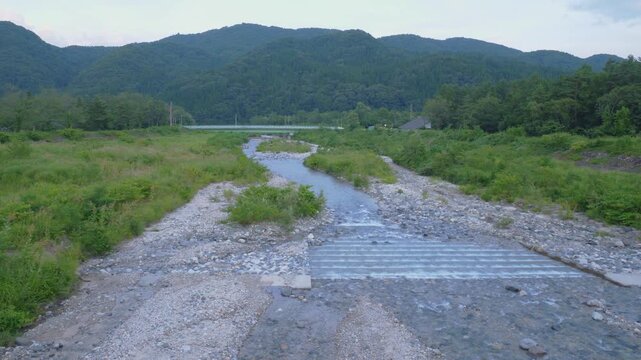 Shallow Rocky River Flowing Through Lush Green Valley With Low Concrete Weir In Nagano, Japan. wide shot