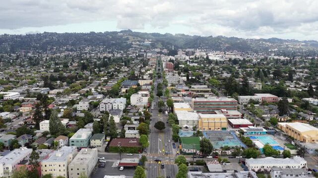 Berkeley tree-lined boulevard stretching toward wooded hills, low-rise residential and commercial blocks spreading across dense urban grid under cloudy sky. California, USA. Aerial rise wide shot.