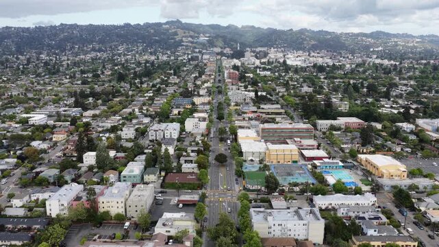 Berkeley wide boulevard stretching toward wooded hills, low-rise residential and school blocks on both sides under cloudy sky with scattered clouds. California, USA. Aerial wide shot. Peaceful town.