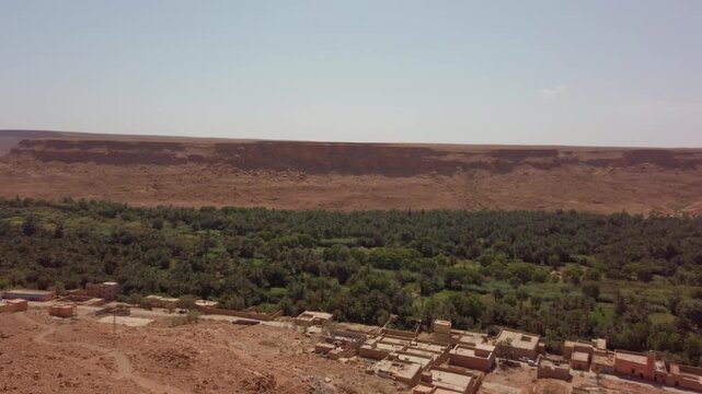 In Morocco's Ziz Valley, an oasis with the Fleuve de Ziz runs through desert terrain bordered by date palms, farmland, and mud brick village structures amid surrounding cliffs.