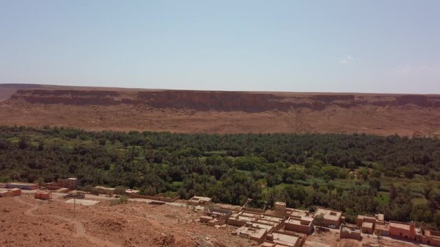 In Morocco's Ziz Valley, a lush oasis with palm trees, greenery and farmland sits beside the Ziz River between desert cliffs and rock formations in a broad panoramic view.
