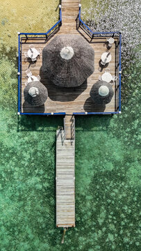 Aerial view of a wooden pier and a deck with thatched umbrellas and white tables over clear turquoise water in George Town, Exuma, The Bahamas.
