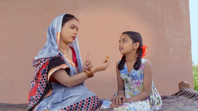 Indian mother feeding her little naughty daughter while sitting on a cot/charpai. Medium shot of a young village woman insisting her cute kid/child to eat some more food - parenting and lifestyle