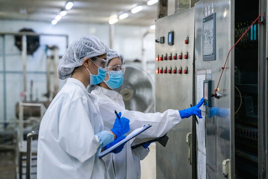 Two individuals wear white lab coats, hairnets, and face masks. One points to a machine while the other holds a clipboard. They use blue gloves during a technical check. The environment is a clean